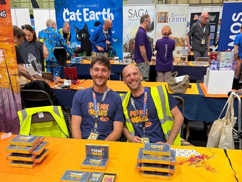 Two men smiling behind a table at a convention booth, wearing matching blue 'Prank Warriors' t-shirts and high-visibility vests. The table displays boxed and loose Prank Warriors cards, with 'Grizth Foundry' and 'SAGA' banners visible in the background.
